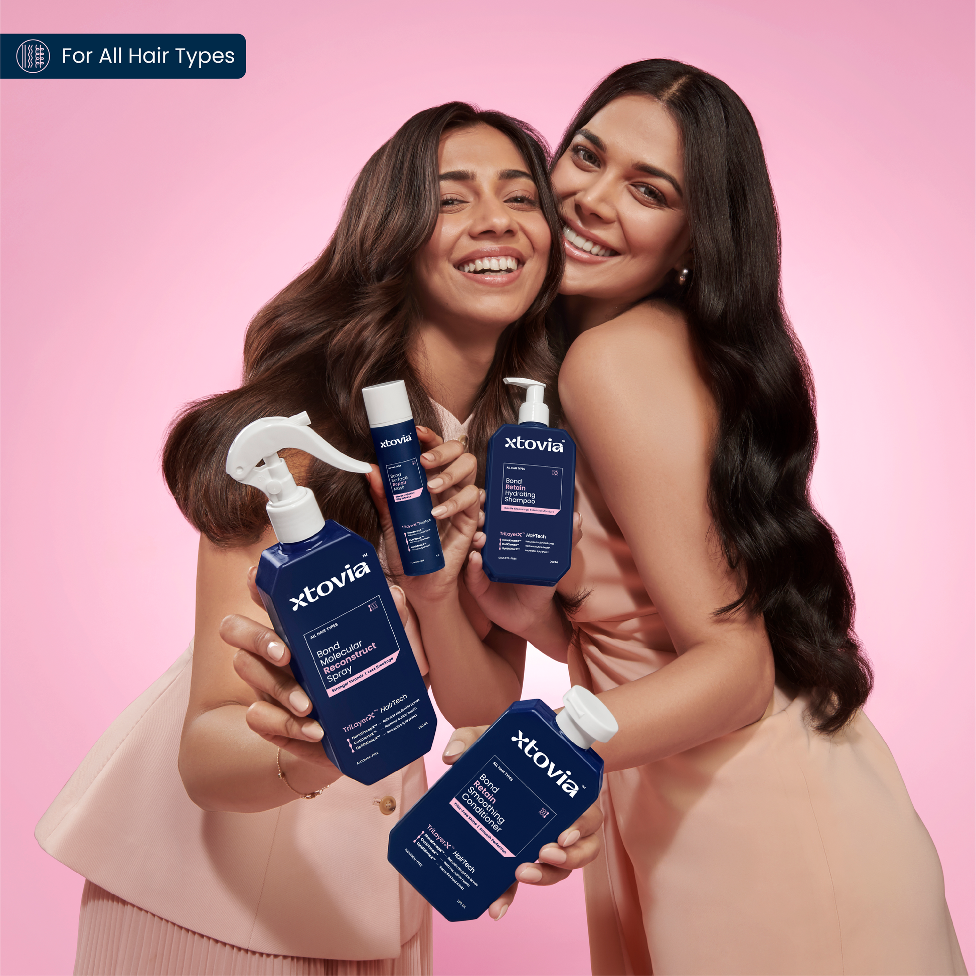 Two women holding Xtovia The Ultimate Hair Revival Kit - Bond Repair Range | Spray, Mask, Shampoo and Conditioner, against a pink background. The image suggests it is for all hair types